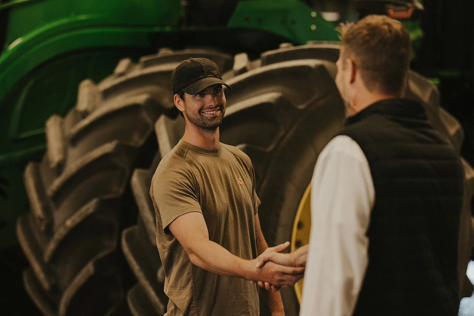 Two men shaking hands in front of large tractor tires, indoor setting.