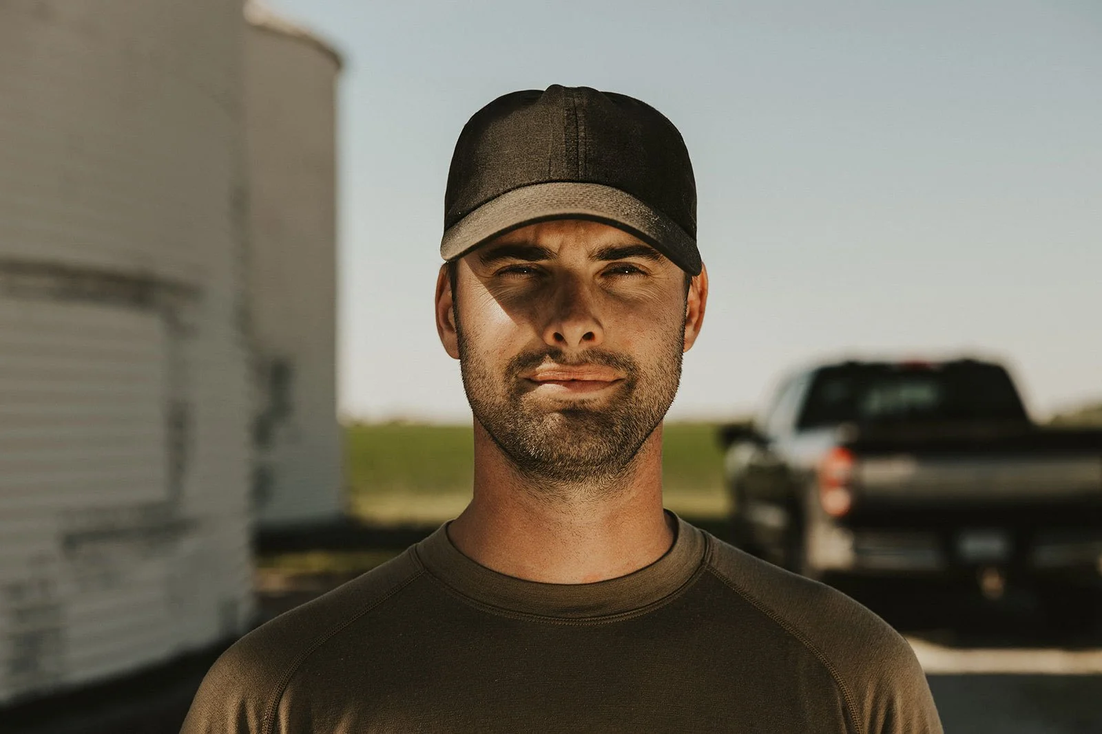 A man wearing a black and beige cap and a dark green shirt, standing outdoors with a blurred background of a black pickup truck and a white trailer.