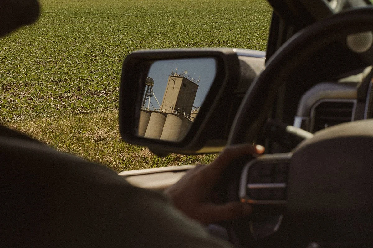 A person driving a vehicle with a farm silo visible in the side mirror.