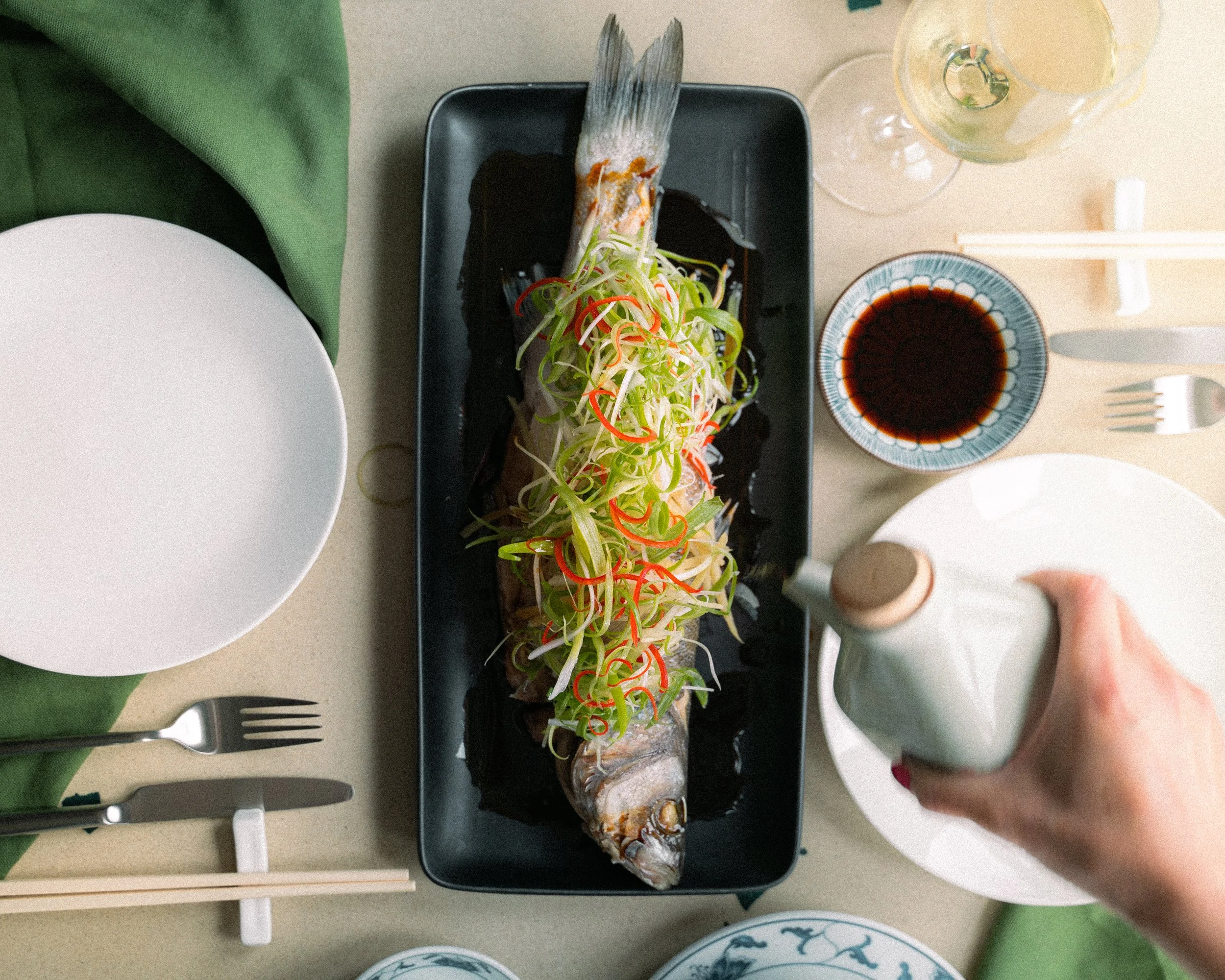 A plate with sushi rolls topped with colorful sliced vegetables and garnished with green onion. The sushi is served on a black rectangular plate, with soy sauce and a drink with lemon wedges on the side. There are empty plates, forks, and a person's hand reaching for the sushi.