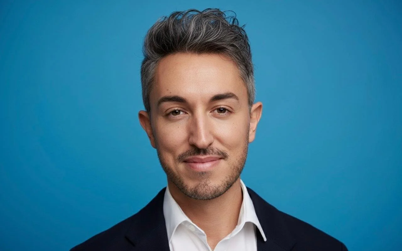 Headshot of a young man with styled grey hair, light facial hair, wearing a white shirt and dark blazer, smiling lightly, against a blue background.