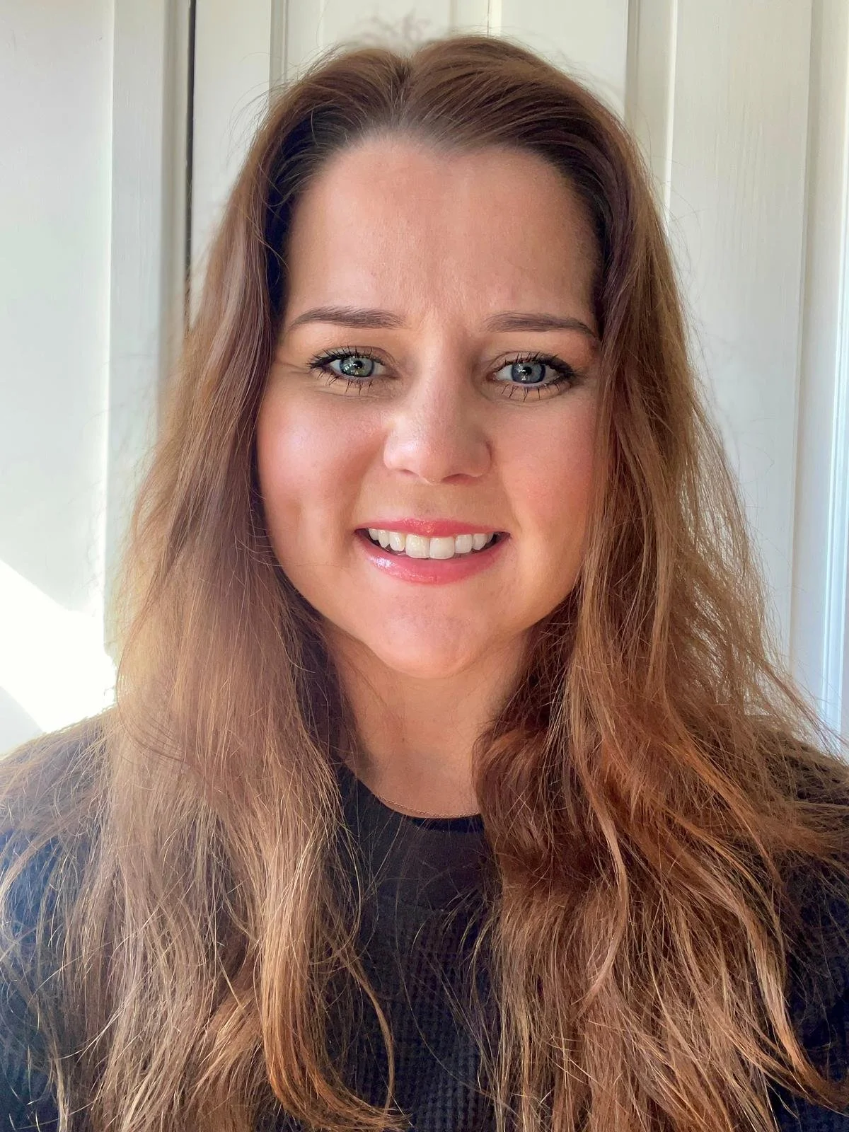 Close-up portrait of a woman with long, wavy brown hair, blue eyes, and light skin, smiling and wearing a black top, standing in front of a white door.
