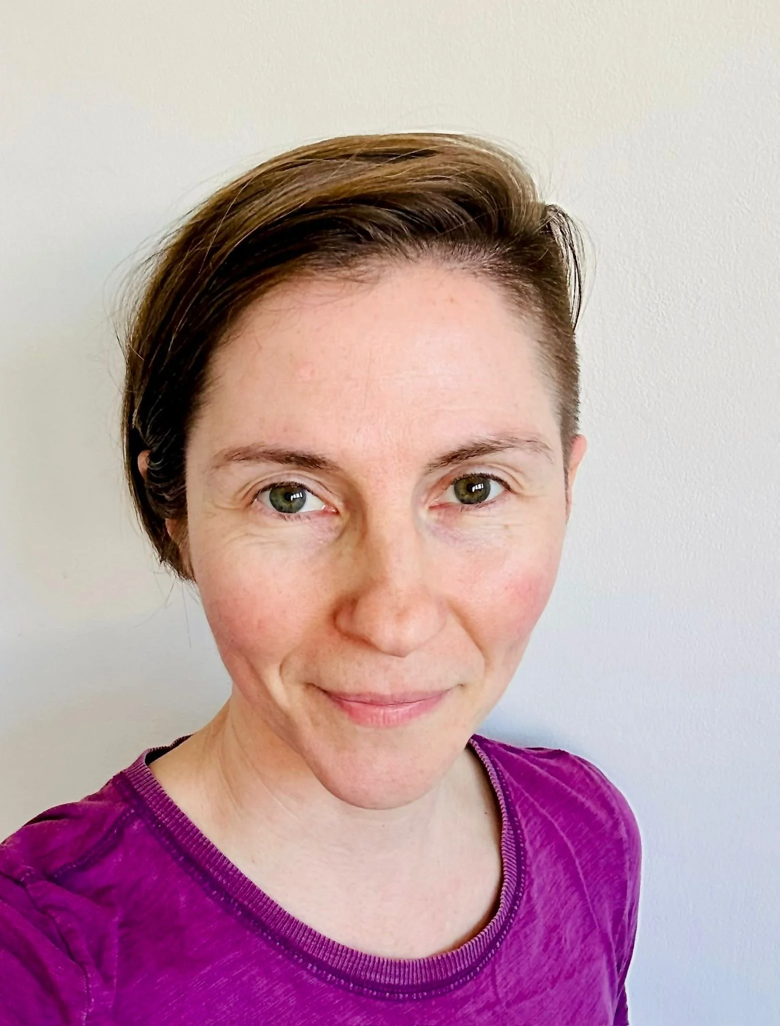 Close-up of a woman with short brown hair and green eyes, wearing a purple shirt, standing against a plain white wall.
