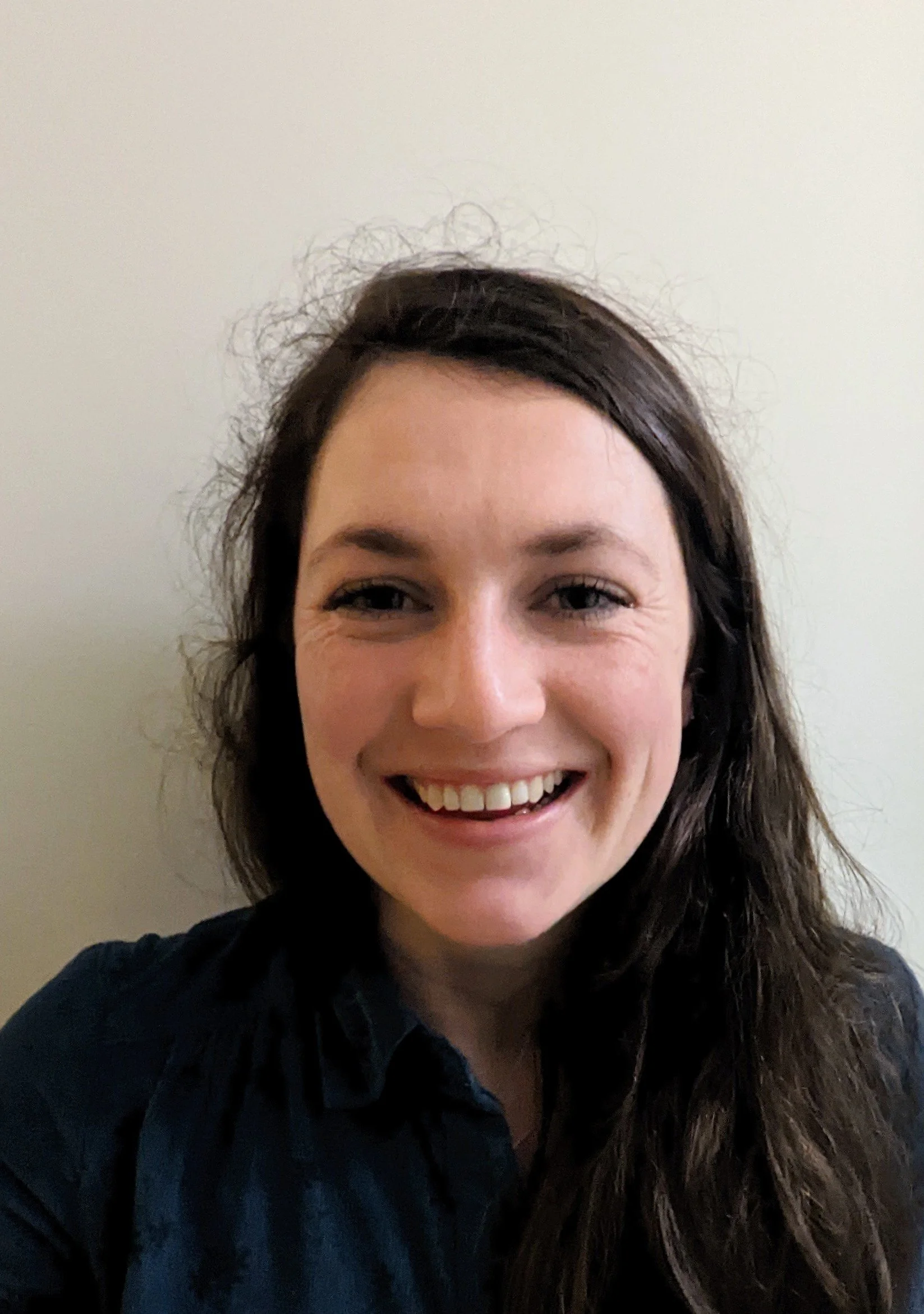 Close-up of a woman's smiling face with long dark hair, wearing a dark top, standing against a plain light-colored wall.