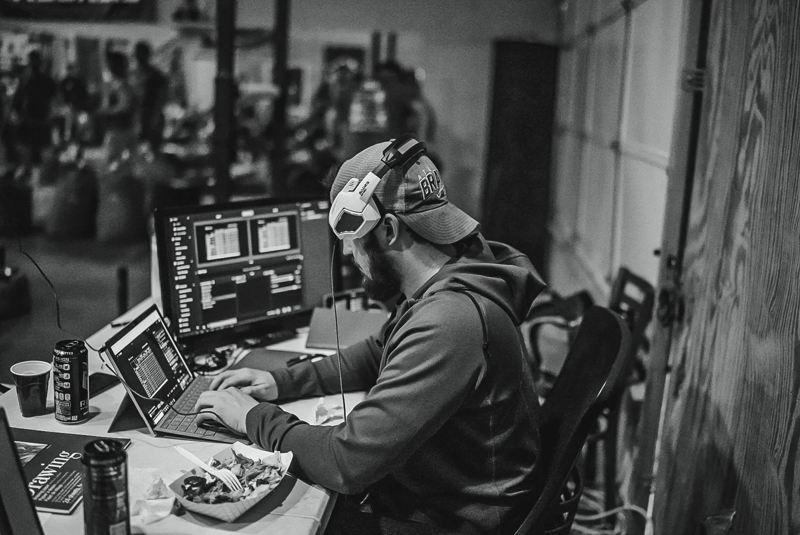 A man wearing a baseball cap and headphones is working on a laptop and two monitors at a table with food and drinks, in what appears to be an indoor setting with a busy, blurred background.