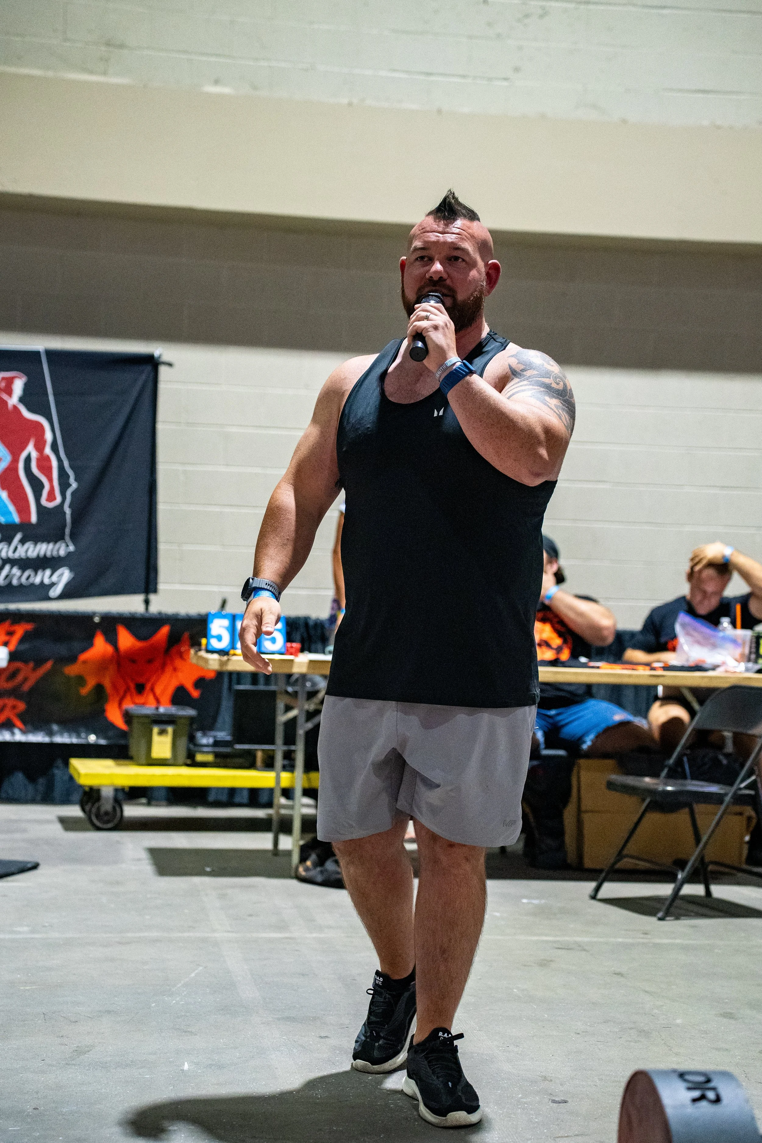 A man with a beard, short hair styled in a mohawk, wearing a black tank top, gray shorts, and black sneakers, speaking into a microphone at an indoor event with a banner, people sitting at tables, and equipment in the background.
