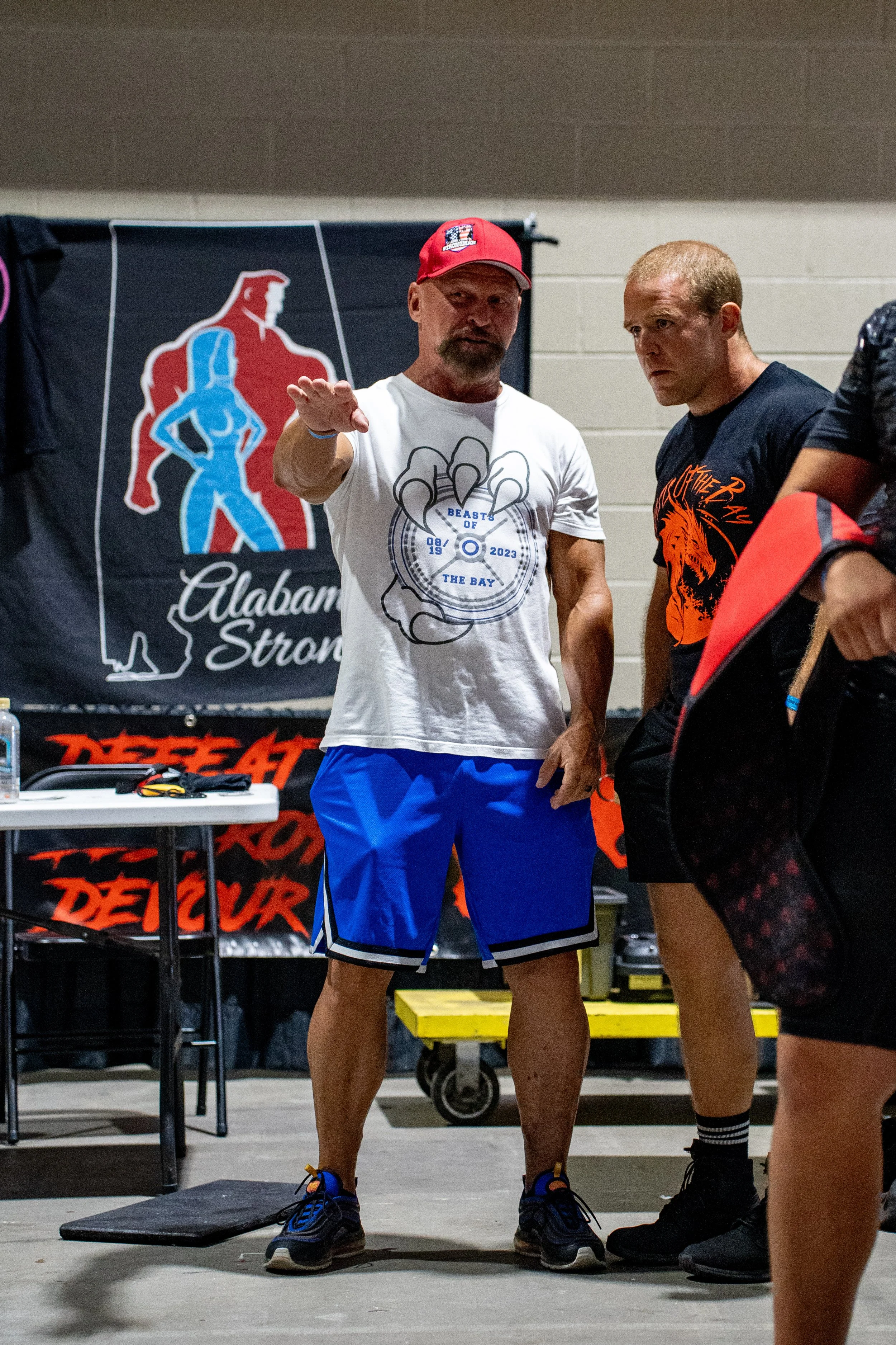Two men talking in a gym or fitness center, with a banner behind them that says 'Alabama Strong'. One man has a beard, wearing a white T-shirt and blue shorts, and the other is shaved-headed wearing a black T-shirt with a lion graphic. They appear to