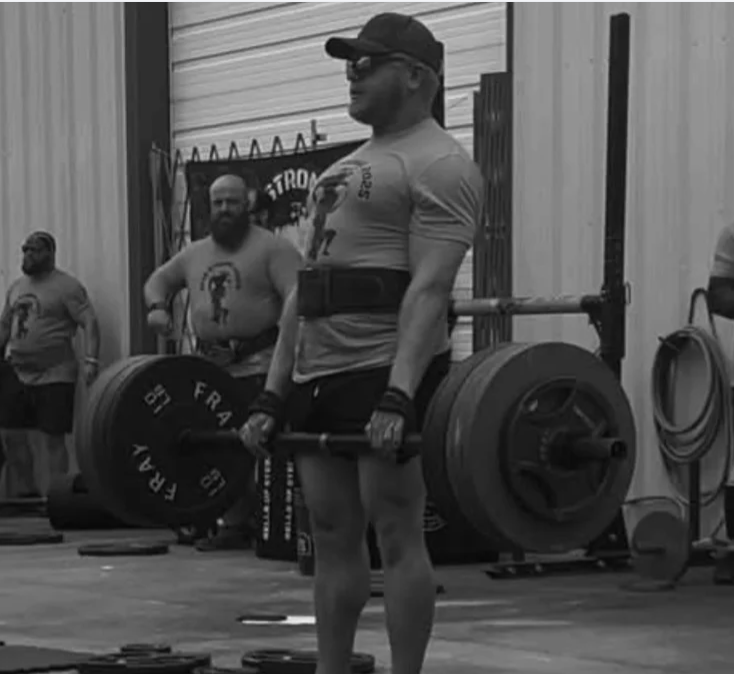 Man lifting heavy barbell at gym, wearing gloves, cap, and sunglasses, with other men in the background.