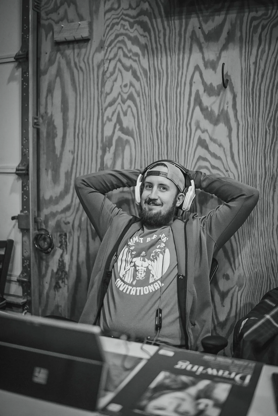 A young man with a beard sitting in a room with wood-paneled walls. He is wearing headphones and a cap, smiling with his hands behind his head. He is dressed in a t-shirt and jacket, with a backpack over his shoulders.