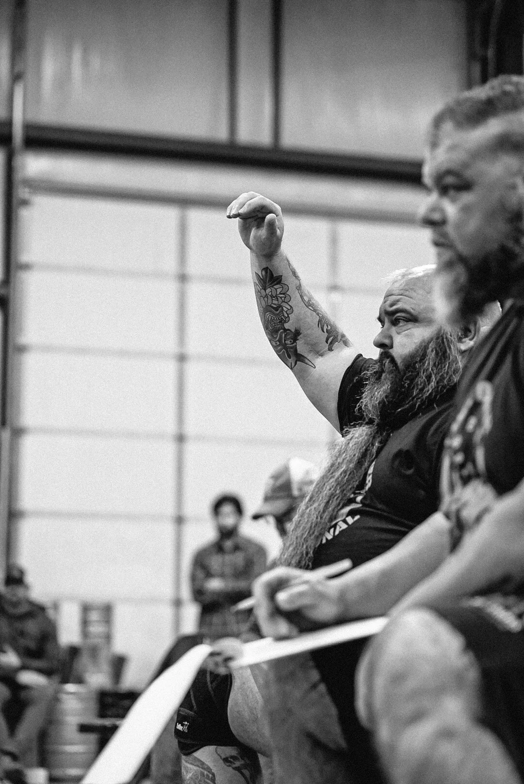 Two men with tattoos sitting outdoors at a bar, one with a beard and long hair, and the other with a buzz cut, both wearing black shirts, engaged in a conversation.