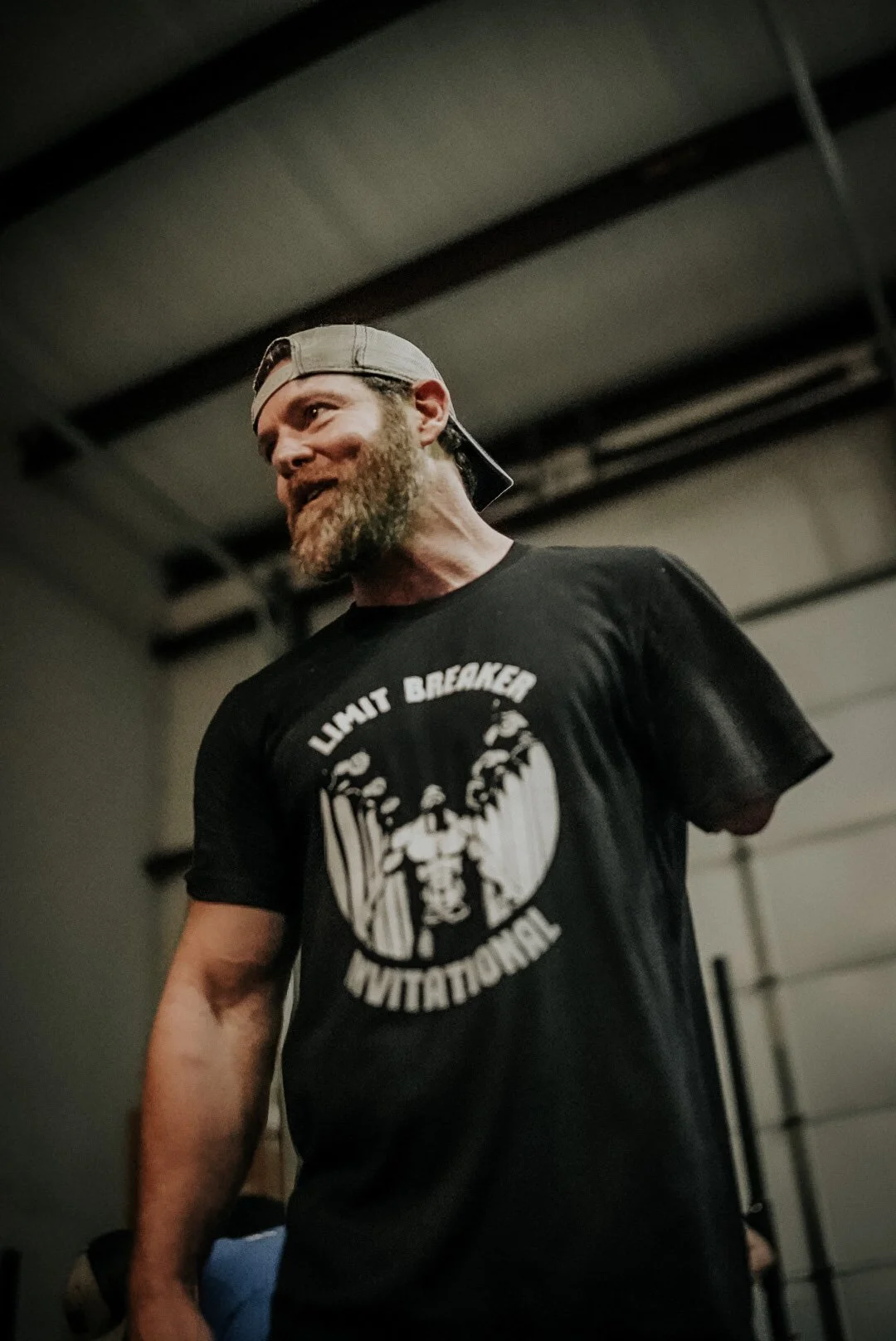 A bearded man wearing a beige cap backwards and a black T-shirt with "Lunat Breaker Invitational" text and a graphic of a fighter in the center.