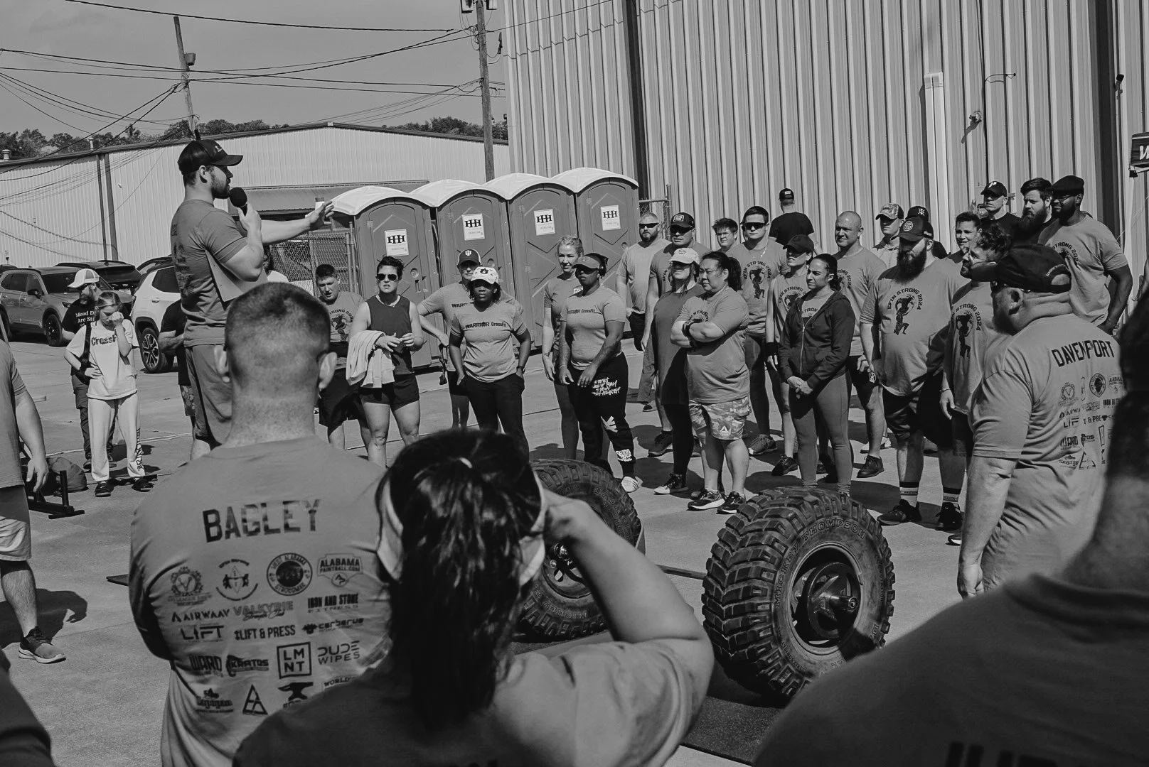 A group of people gathered outdoors, listening to a man speaking with a microphone, with large tires in the foreground.
