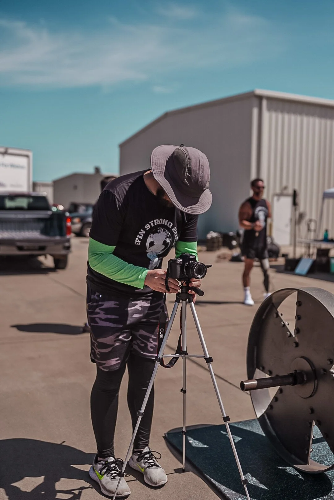 A person wearing a wide-brimmed hat and a black shirt with green sleeves is adjusting a camera on a tripod outside, with another person in the background near a building.