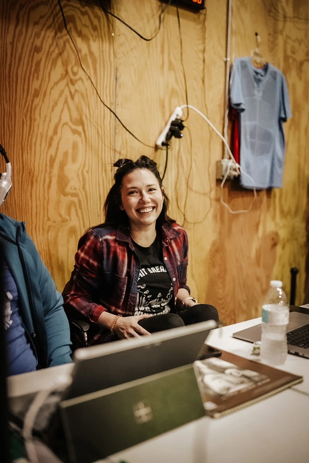 A young woman with dark hair smiling while sitting at a table in a room with wooden walls. She is wearing a black T-shirt with a graphic and a red plaid shirt. There are laptops, a water bottle, and some books on the table. A blue shirt is hanging on