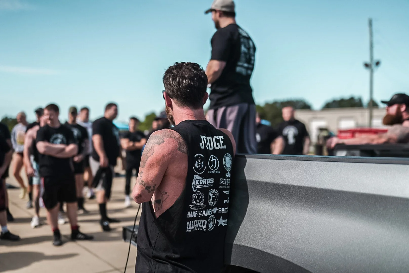 A muscular man with tattoos and a sleeveless black shirt labeled 'JUDGE' in a group of people outdoors under a clear blue sky, with some standing and one sitting on a truck tailgate, at a fitness or strength event.