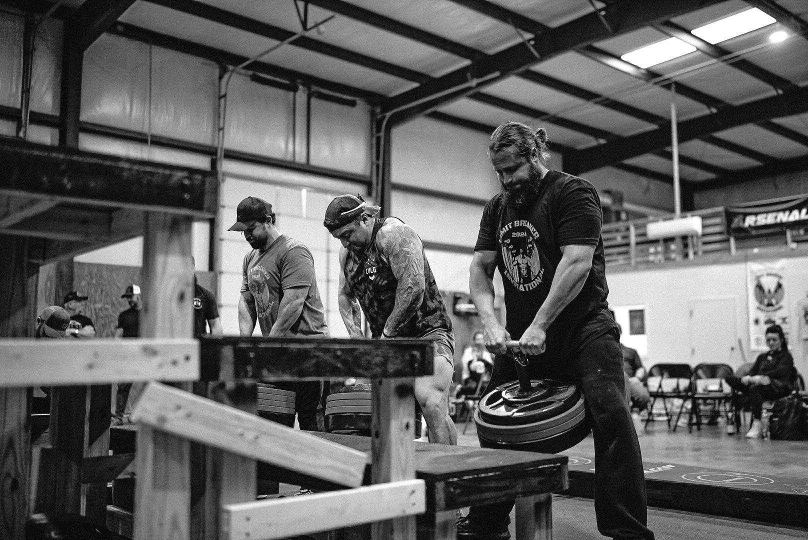 Three men working in a gym with weights, one man is lifting a weight plate, two men are standing and preparing for exercise, equipment and chairs are visible in the background.