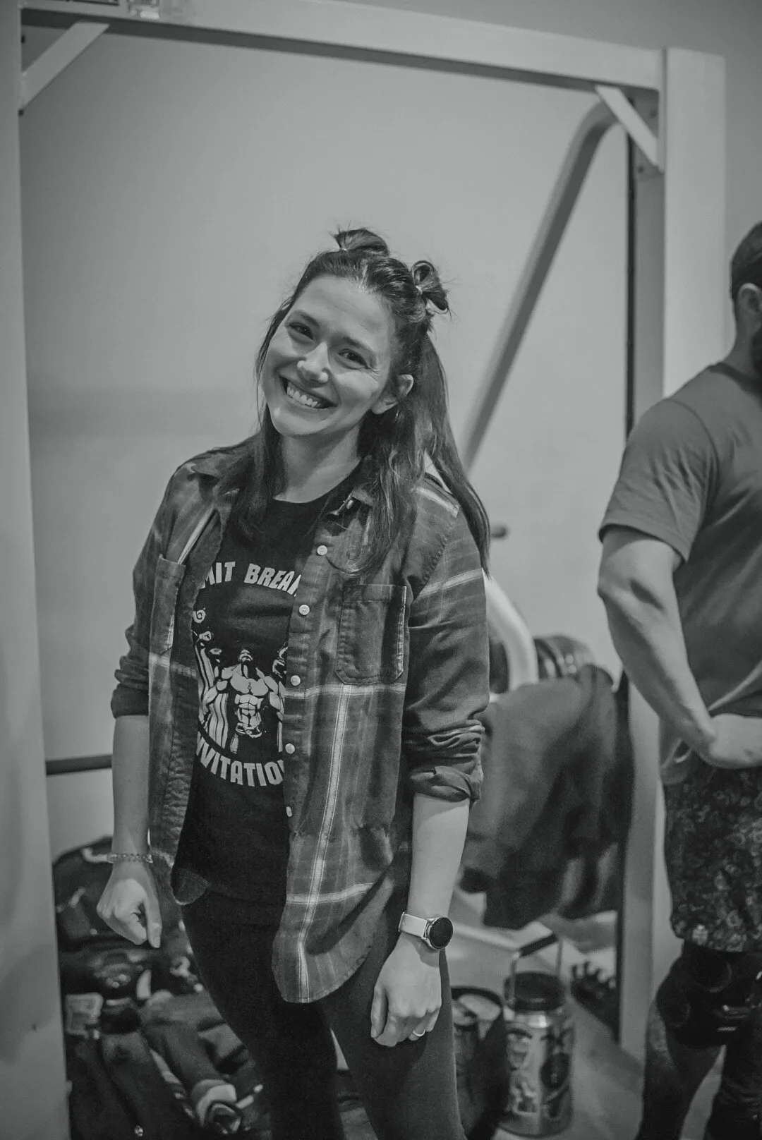 Smiling woman with wavy hair tied up in a bun, wearing a plaid shirt over a graphic t-shirt, standing indoors with bags and other items in the background.