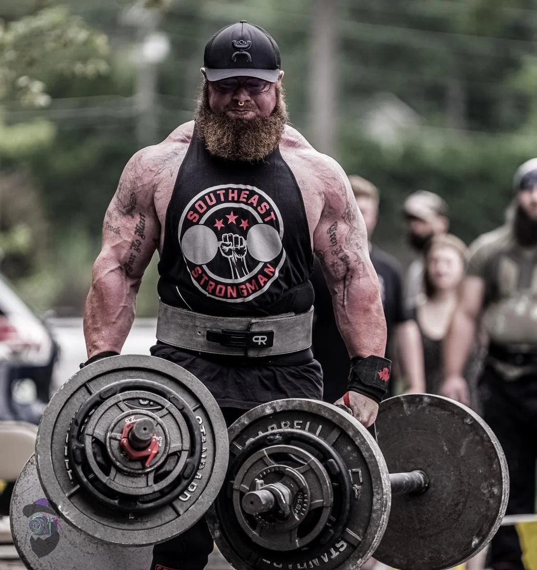 Man with a beard lifting weights outdoors, wearing a black cap and tank top with "Southeast Strongman" logo, in front of a group of people.