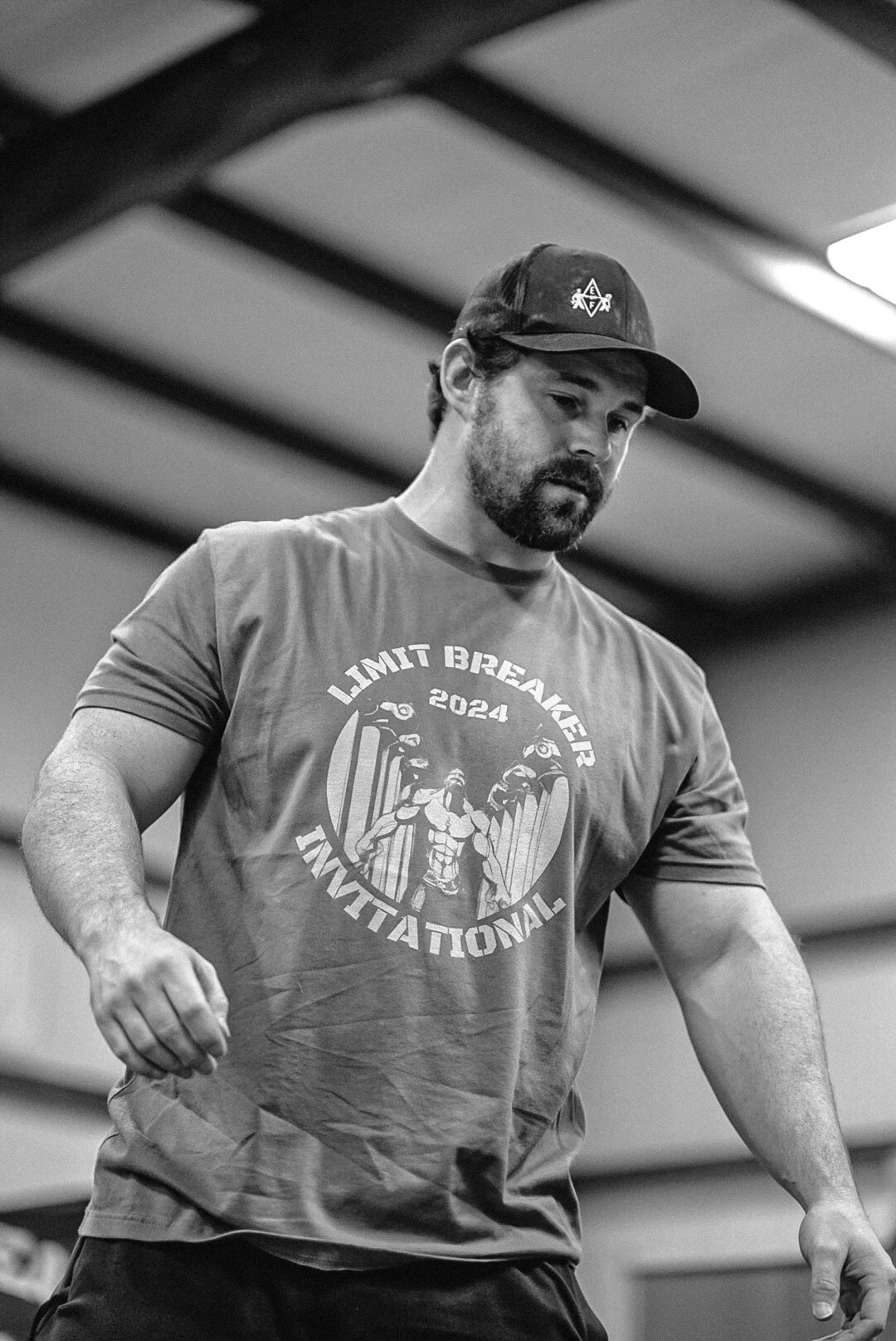 A man with a beard and cap working out indoors. He is wearing a T-shirt that reads "Limit Breaker 2024 International" with a graphic of a muscular figure lifting weights.