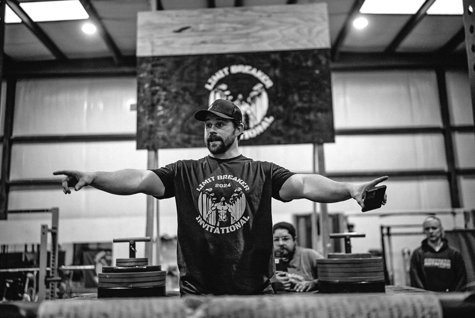 A man standing in a gym, pointing to the left, wearing a cap and a T-shirt that reads "Limit Breaker Invitational 2024" with a muscular figure design, in front of a blurred background with a large banner.