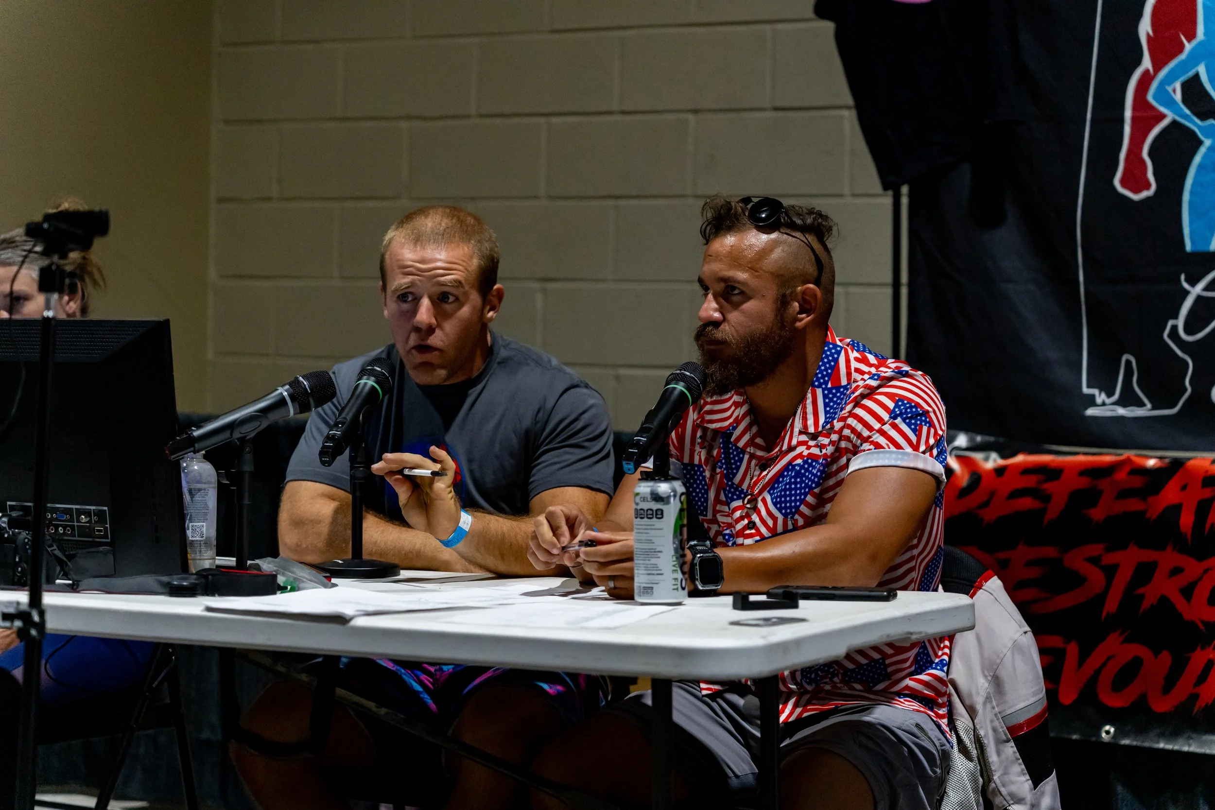 Two men sitting at a table speaking into microphones during a panel discussion, with computer equipment and beverage bottles in front of them.