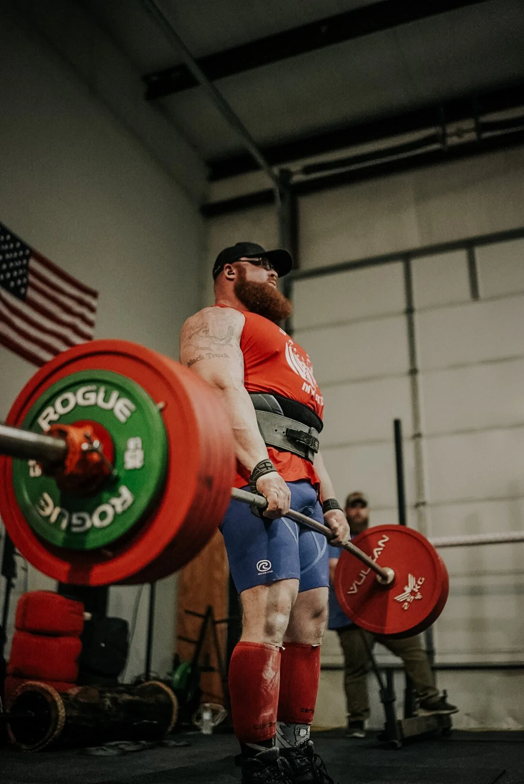 A muscular man with a beard, wearing a red shirt, blue shorts, and red knee-high socks, is lifting a barbell with red and green weight plates in a gym. An American flag is visible in the background.