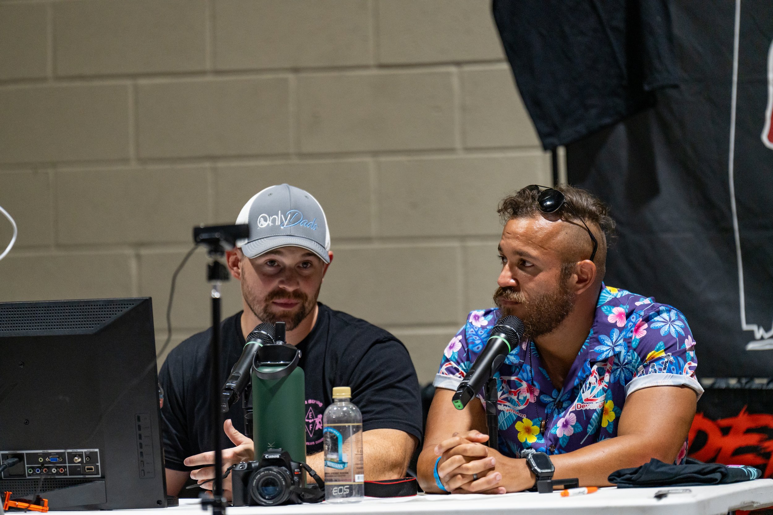 Two men are sitting at a desk with microphones, computer, and water bottles, engaged in a discussion or podcast. The man on the left is wearing a gray baseball cap with the words '@Dad' on it, and the man on the right is wearing a colorful Hawaiian s