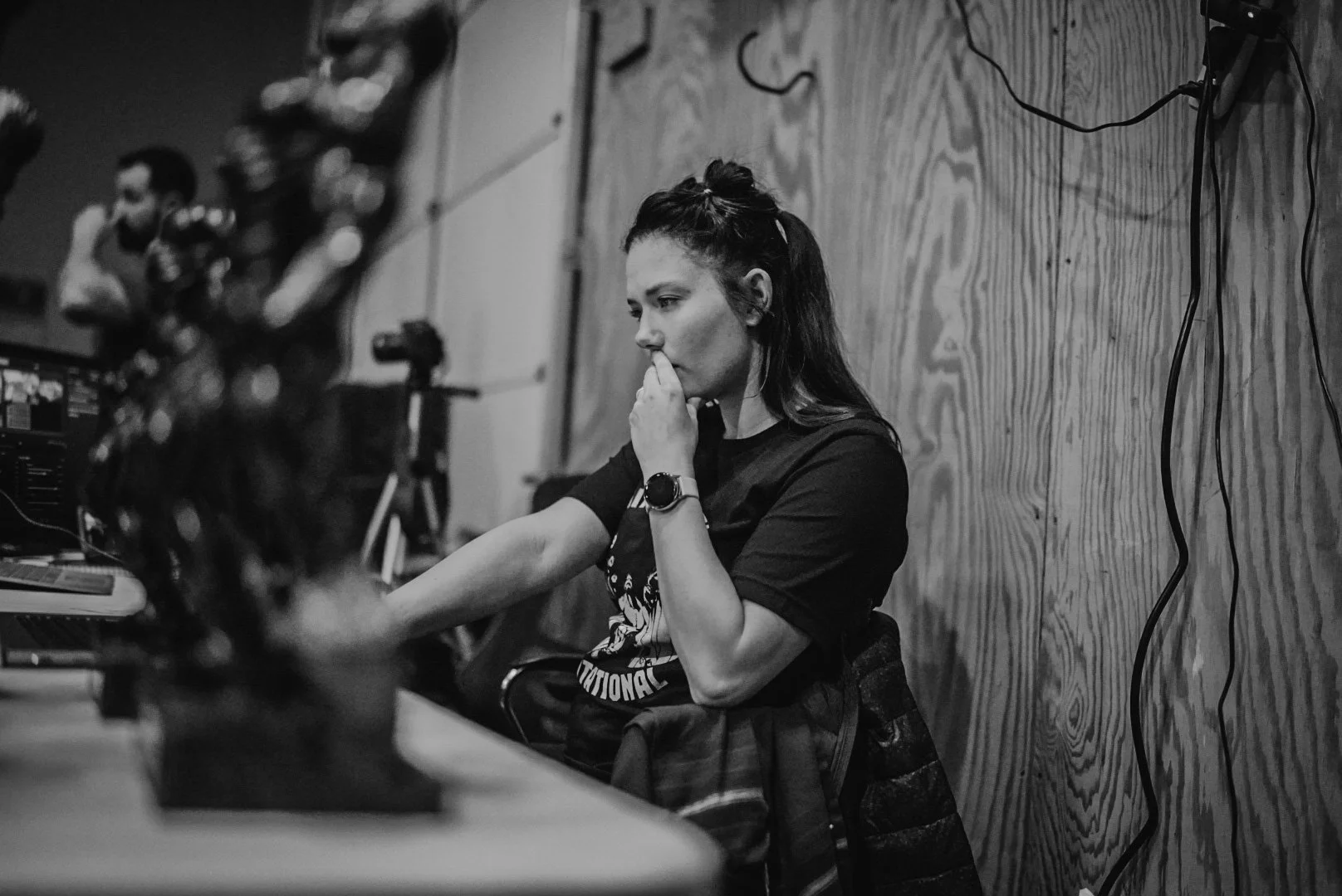 A woman with long hair tied back, wearing a black t-shirt and a watch, sitting at a desk with her hand on her chin in a contemplative pose in a room with a wooden wall and visible cables.