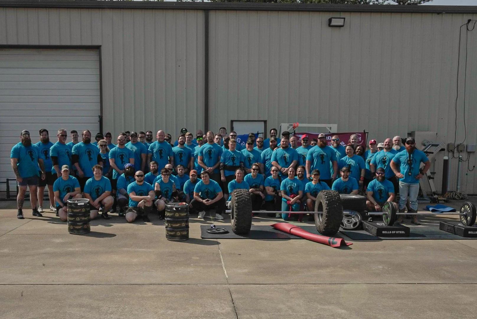 Group of people wearing matching blue shirts assembled outside a building with gym equipment and weighted barrels in front.