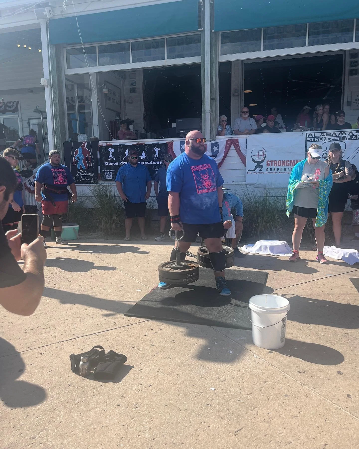 A man with a beard, sunglasses, and a blue T-shirt stands on a black mat, lifting weights outdoors at a strength competition. Several other people are in the background, some observing and others preparing. There are banners and signs related to stre