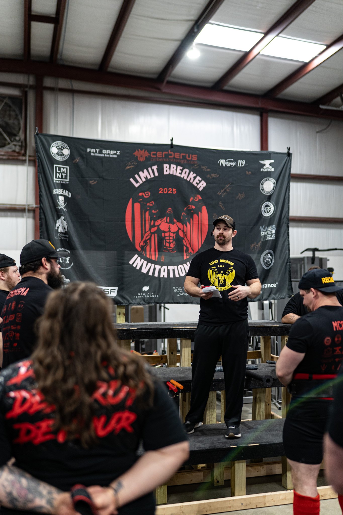 A man wearing a black cap and black T-shirt with yellow graphics and text stands in front of a large banner at a strength competition, with several people seated and standing around him.