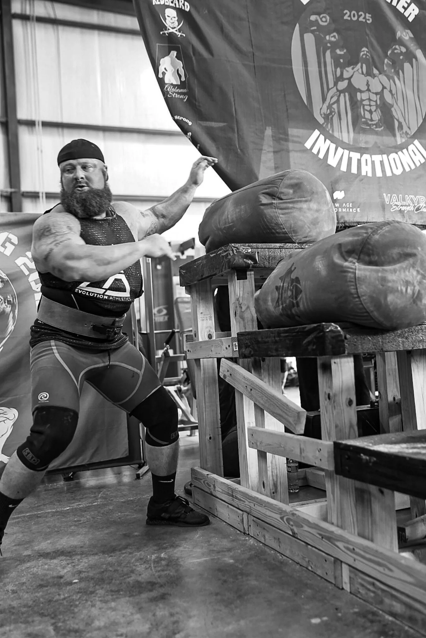 A muscular man with a long beard, wearing a black headband and athletic clothing, is in a gym setting and appears to be participating in a strongman competition or training. He is lifting a wooden frame that holds large, round bags, which are likely 