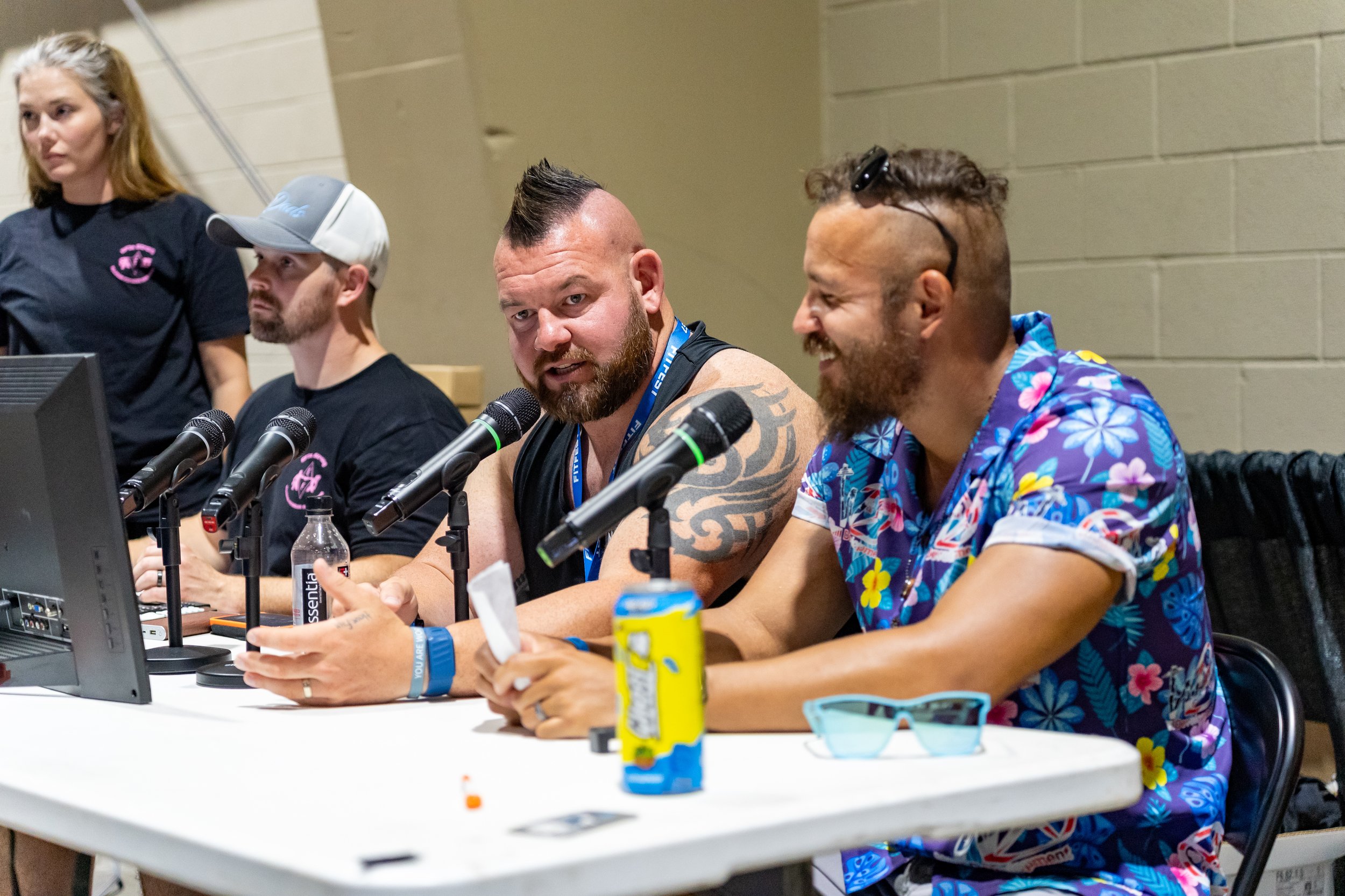 A panel of four people sitting at a conference table with microphones, water bottles, and snacks, engaged in discussion. A woman stands in the background, and the speakers are dressed casually, some wearing festive, colorful shirts.
