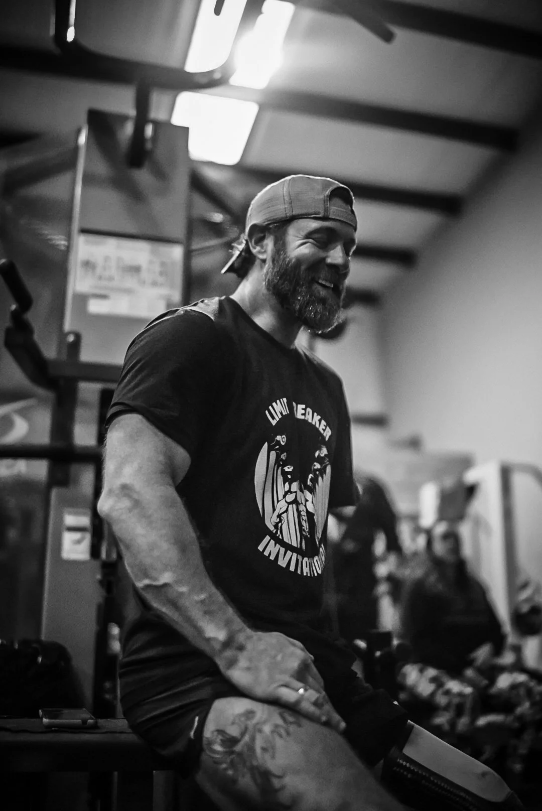A man with a beard and tattooed arm, wearing a backwards baseball cap and a T-shirt, smiling and sitting in a gym.
