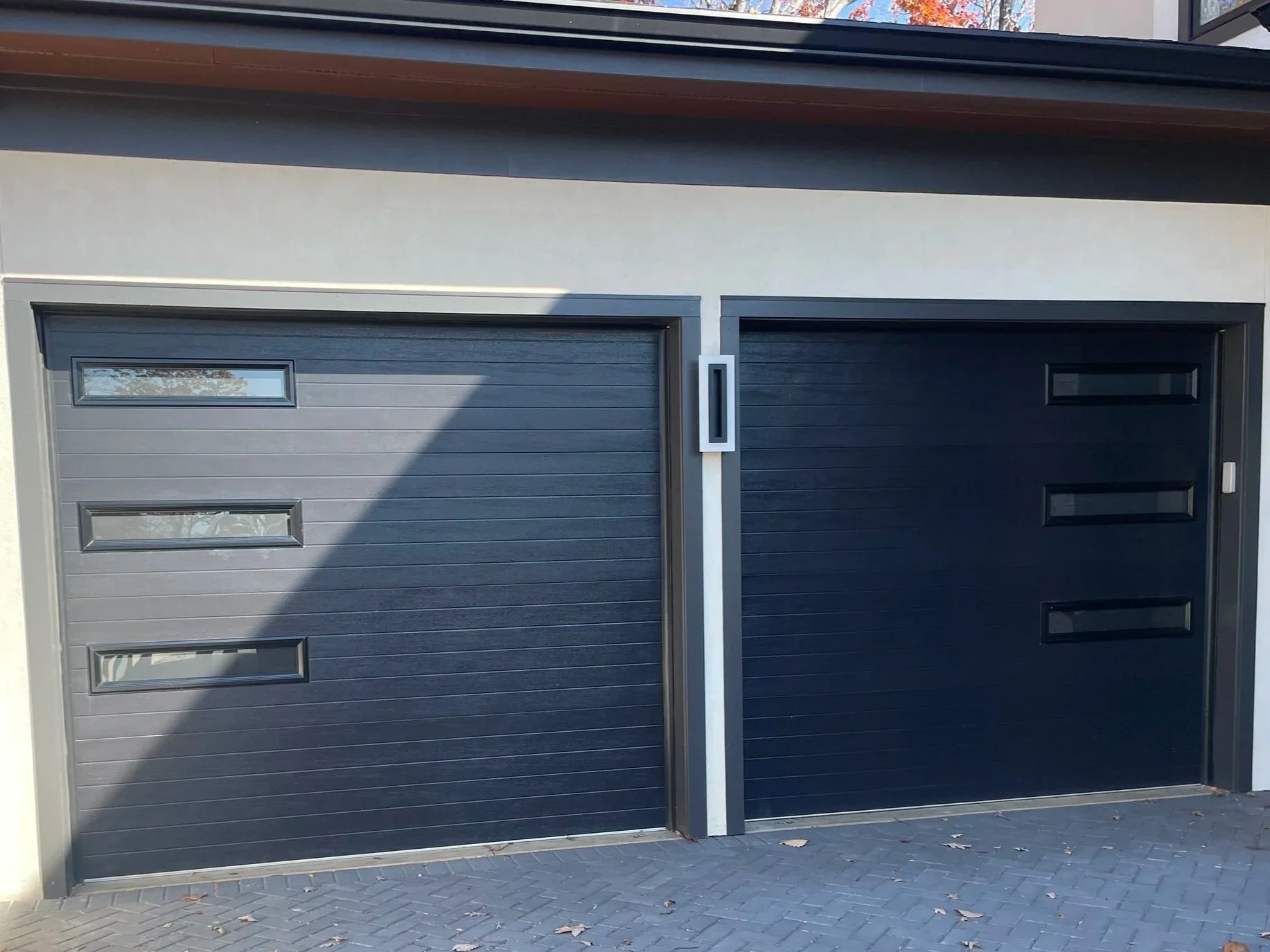 Two modern garage doors, both black, with rectangular windows, set in a contemporary house with a paved driveway.