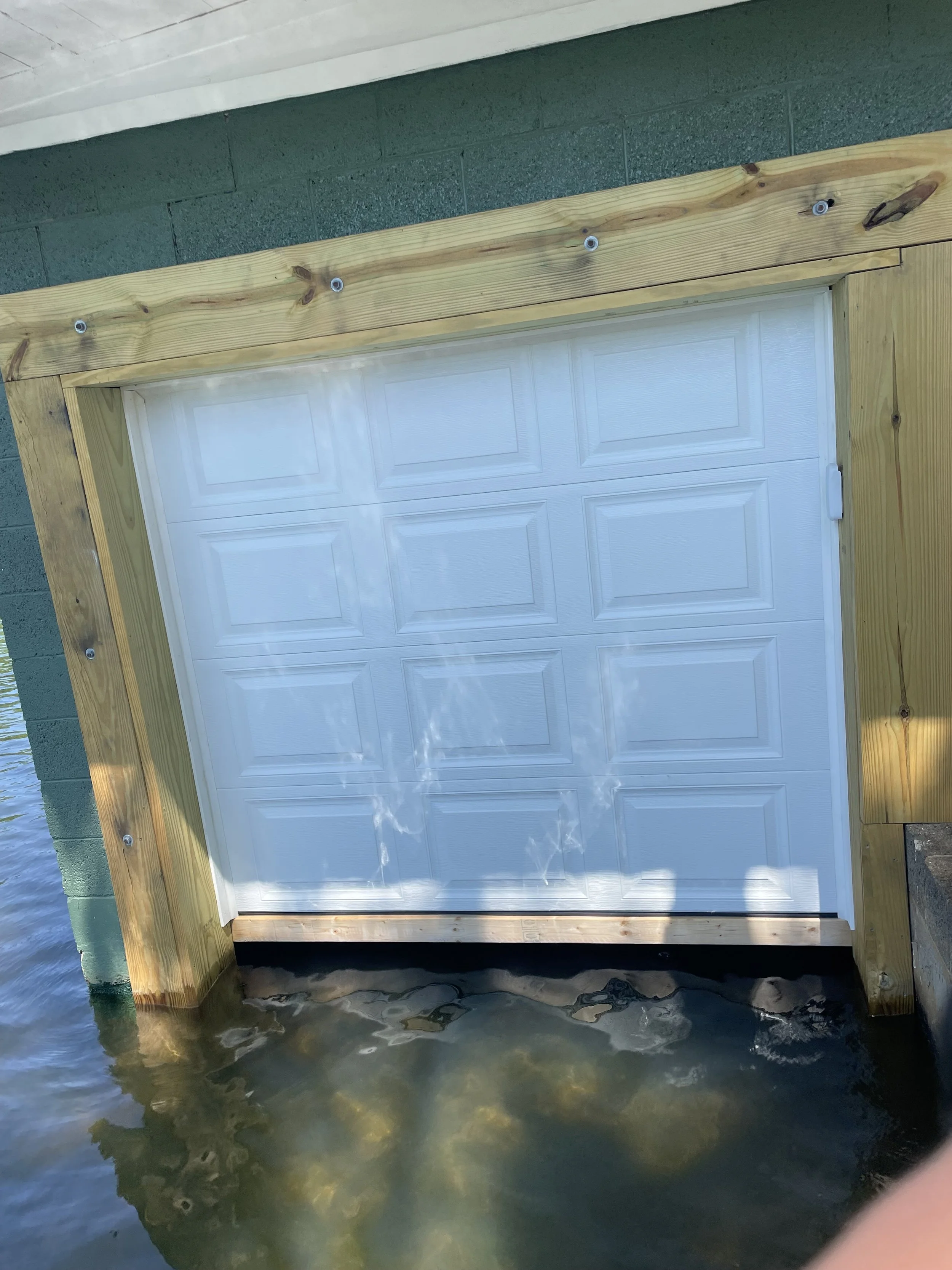 A white garage door installed on a structure located over water, with a wooden frame around it, and sunlight reflecting on the water's surface.