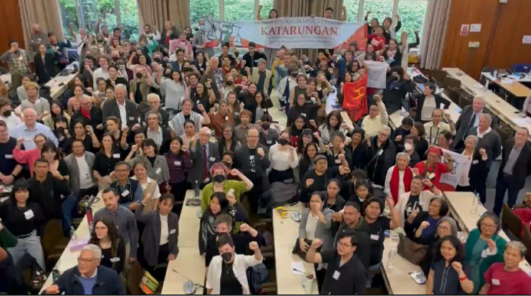 Large group of people attending a conference or seminar in a wooden-paneled hall, with a banner above that reads 'KATARUNGAN.' Some individuals are raising their fists, and the room is filled with tables, chairs, and attendees.