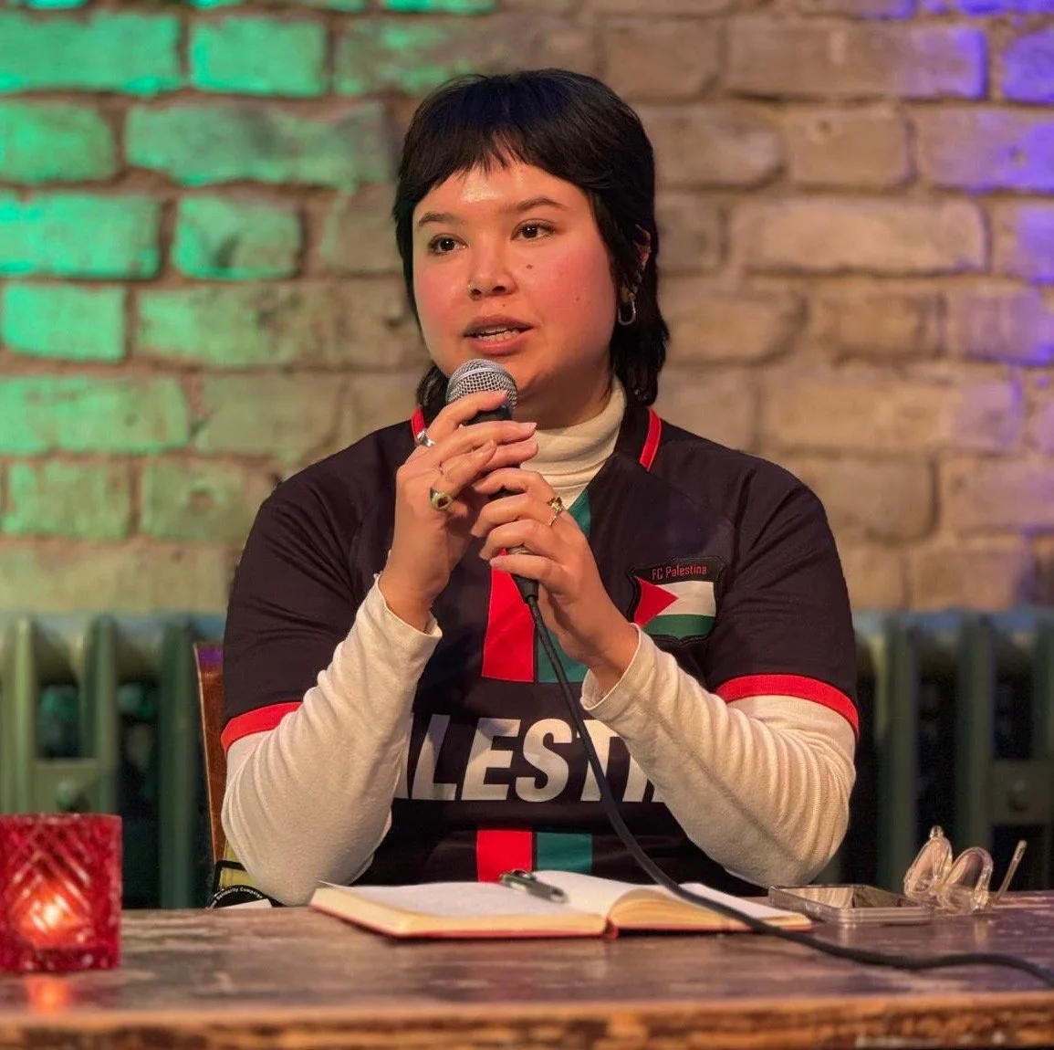 A woman speaking into a microphone at a table with an open notebook, wearing a black and cream jacket with red and green accents, seated in front of a brick wall with colored lighting.