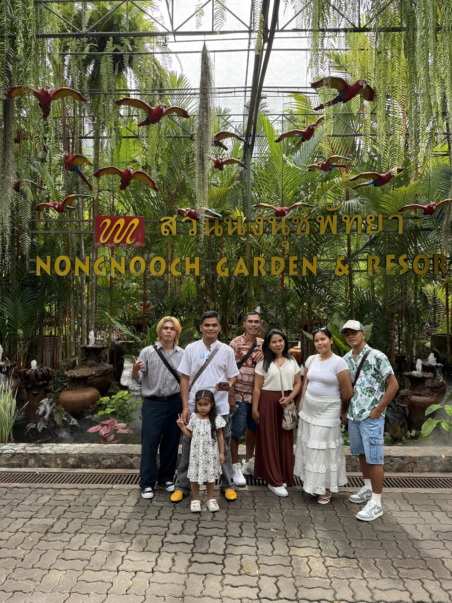 Group of people standing in front of lush green plants inside Nongnooch Garden & Resort, including five adults and one child, with a sign overhead displaying the garden's name in Thai and English.