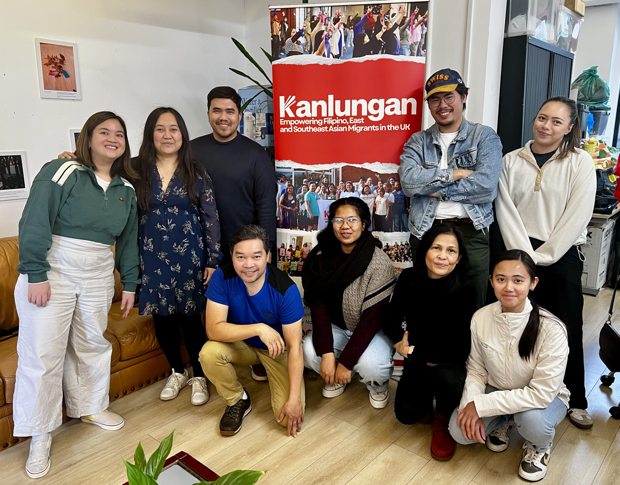 Group of diverse Filipino, East, and Southeast Asian migrants smiling and posing for a photo in front of a banner that says "Kanlungan," indicating empowerment of Filipino, East, and Southeast Asian migrants in the UK. The group includes men and women of various ages, some standing and some sitting, in an indoor setting with posters and shelves around.
