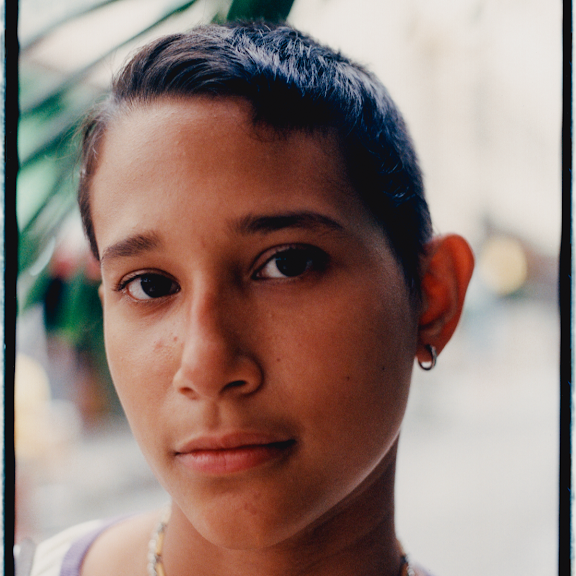 Close-up of young person with dark hair, light skin, and earring, looking at the camera with a neutral expression, outdoors with blurred background.