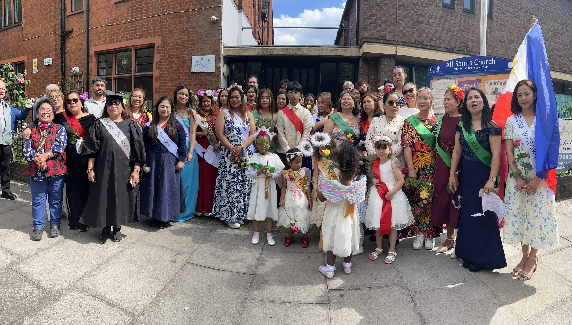 A diverse group of people, including children and adults, gathered outdoors in front of a brick building, some wearing sashes, costumes, and flower crowns, participating in a community or cultural event.