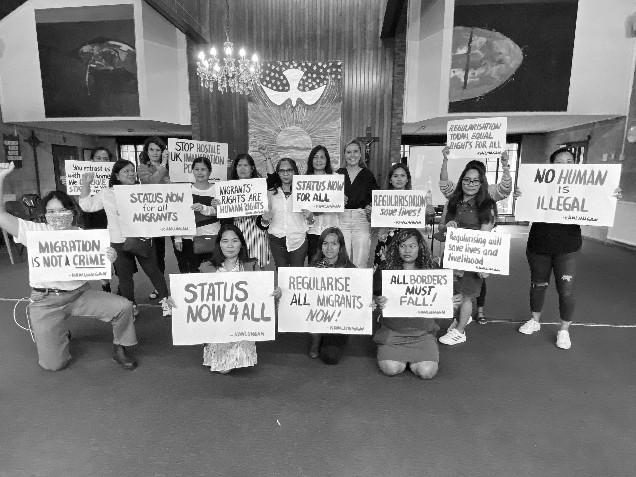 Group of people holding protest signs advocating for migrant rights, regularization, and immigration reform inside a church or community center with a crucifix and religious artwork in the background.