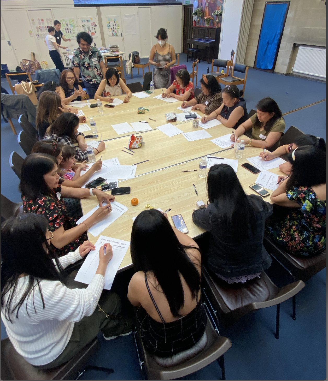 A group of women seated around a large rectangular table, engaged in a meeting or workshop, with papers, pens, and water bottles on the table. Some women are writing, while others are listening or looking at their phones. In the background, a few individuals are standing and organizing materials in a room with colorful decorations and a piano.