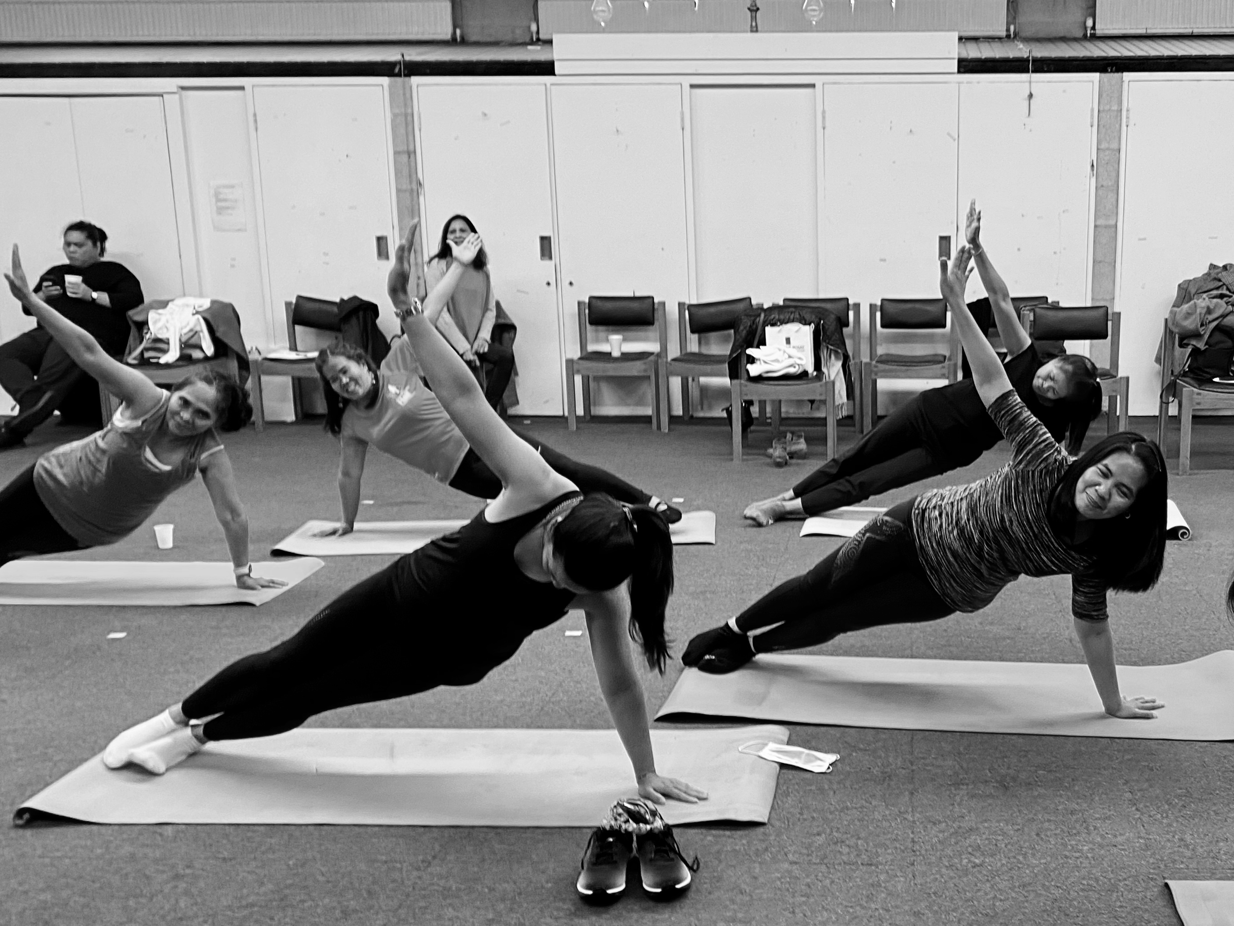 Several women practicing yoga in a studio, performing side plank poses on yoga mats. Some women are raising their arms, smiling and looking at the camera. Chairs and jackets are visible in the background.