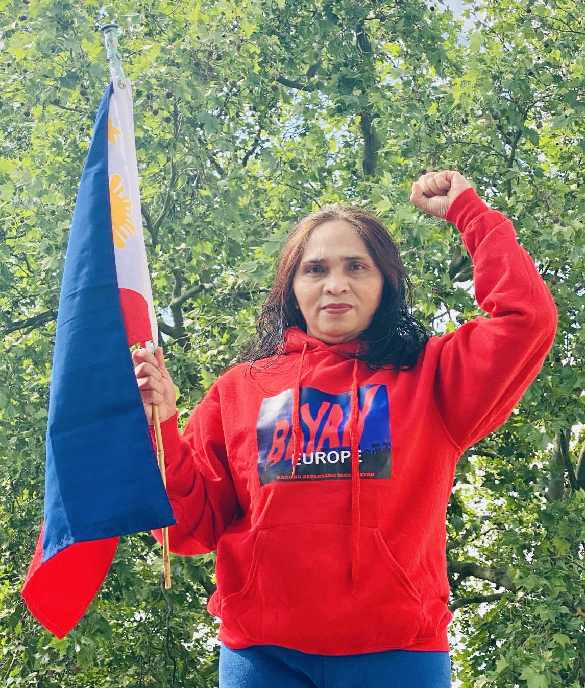 Woman wearing a red hoodie with a political slogan, holding a flag and flexing her arm in a gesture of strength, standing outdoors against a backdrop of green foliage.