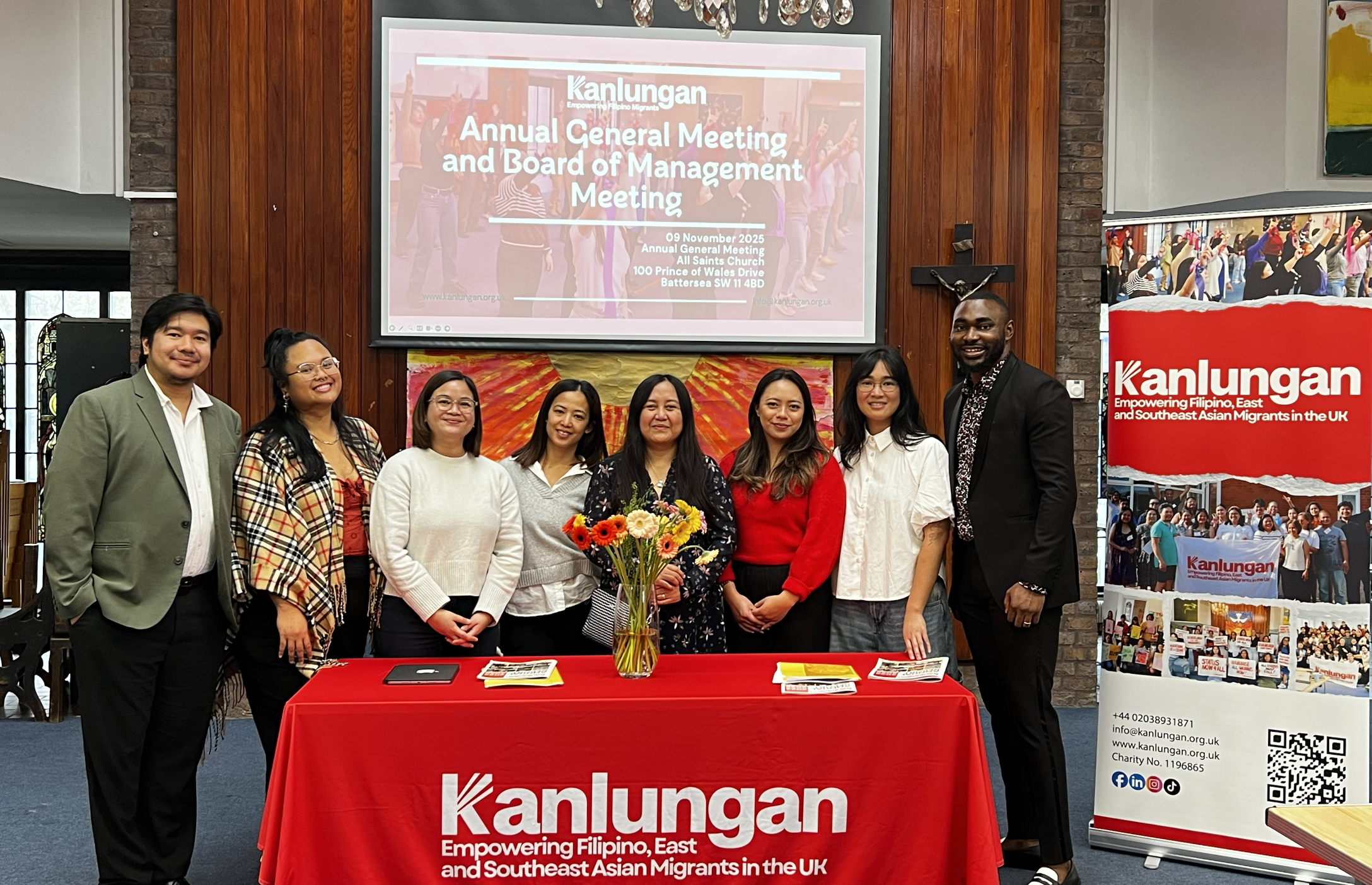 A group of nine people standing behind a red table with a bouquet of flowers on it at an indoor event. Behind them, a large screen displays information about the Kanlungan Annual General Meeting and Board of Management Meeting held on November 9, 2025, at All Saints Church in Battersea, UK. The table and the screen are set against a backdrop of wooden paneling and brick walls. There are colorful posters and banners related to Kanlungan, an organization that supports Filipino, East, and Southeast Asian migrants in the UK.