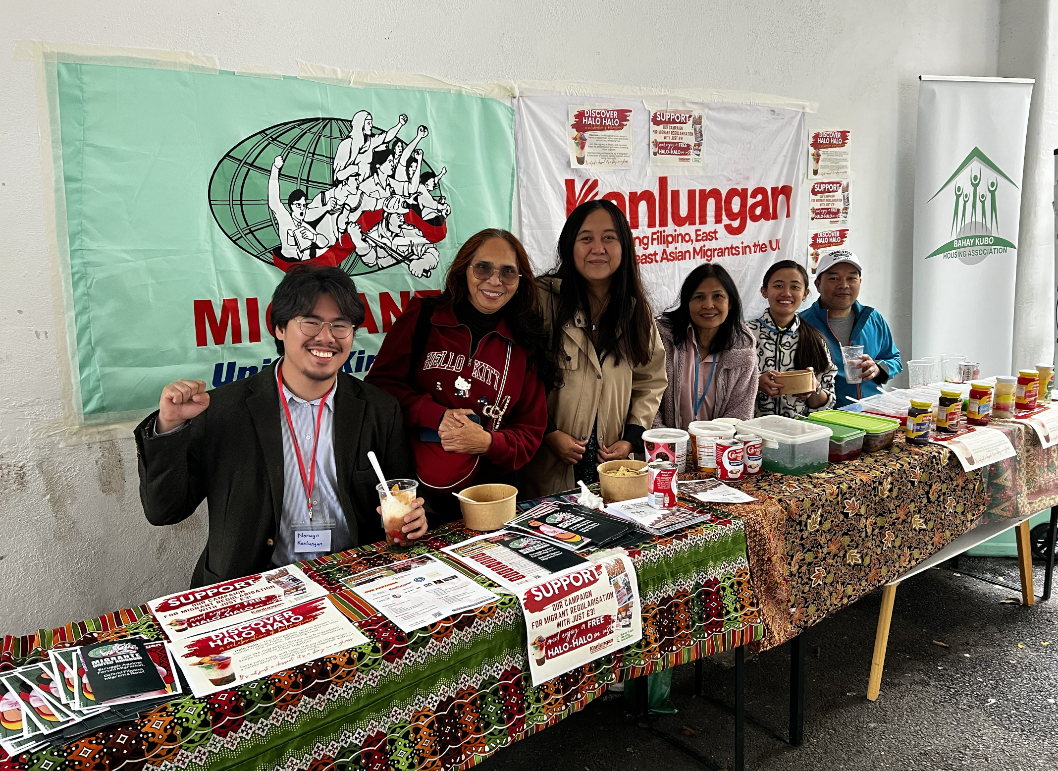 Group of people standing behind a table at a community event, with banners related to migrant support and housing organizations in the background, and various food items and flyers on the table.