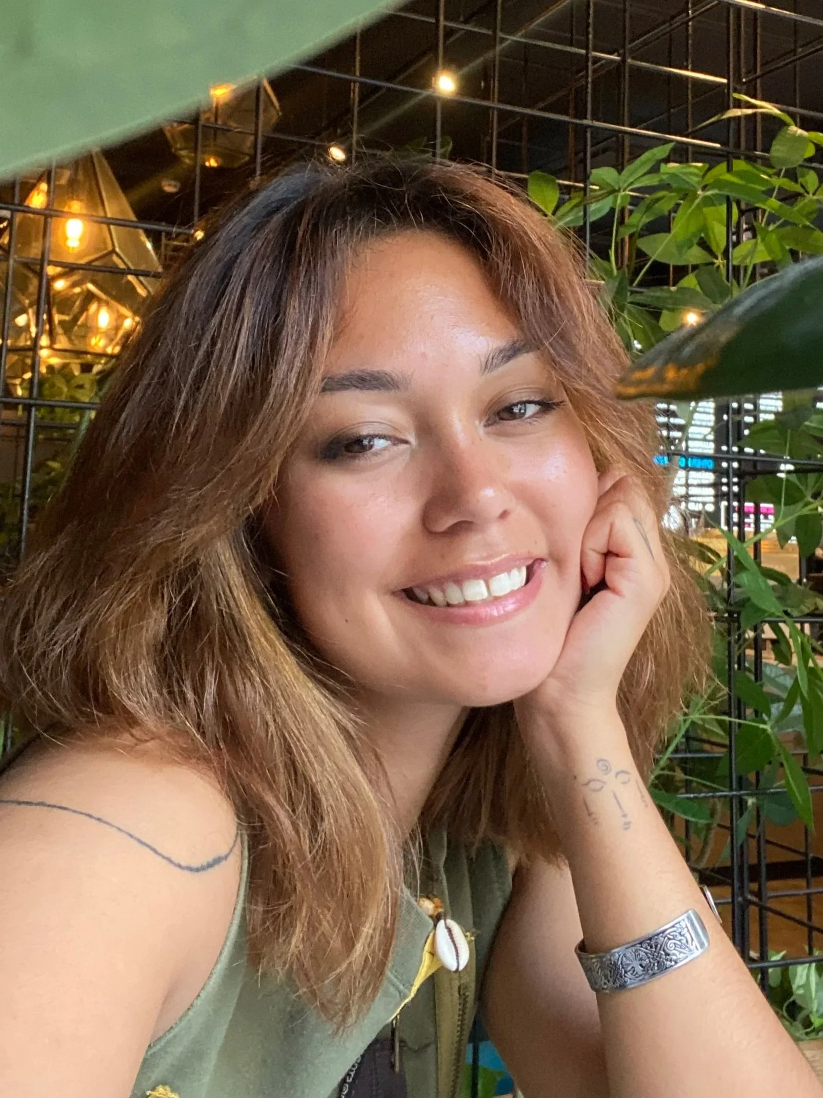 Close-up of a smiling woman with long, wavy, light brown hair, sitting in front of a black wire grid with green plants, warm lighting, and a blurred background in an indoor setting.