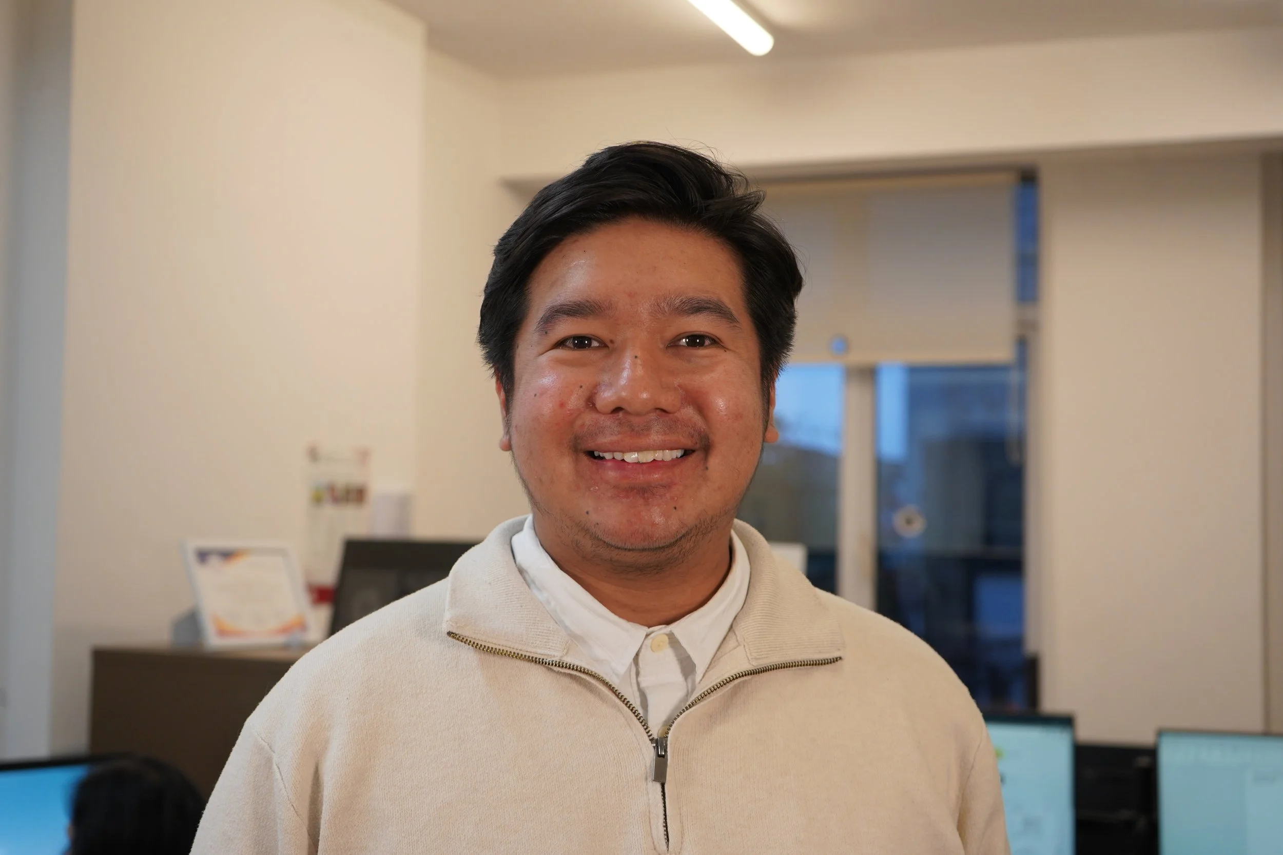 A smiling man with dark hair wearing a light-colored zip-up jacket in an indoor setting with a computer and monitor in the background.